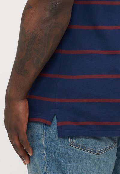 Navyblauwe polo met rode horizontale strepen, textuurstof, zijsplitten aan de zoom, gecombineerd met lichtblauwe denim jeans.