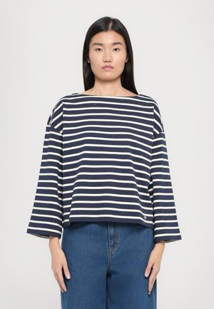 Marineblauw en witte gestreepte, losvallende top met een boothals, wijde mouwen en een cropped ontwerp. Gecombineerd met blauwe jeans.