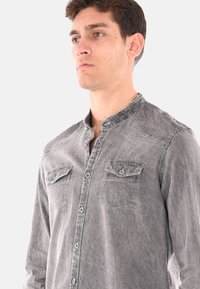 Camicia grigia a maniche lunghe in morbido denim sbiadito, con due tasche sul petto, pattina con bottoni e colletto arrotondato, caratterizzata da dettagli di cucitura visibili.