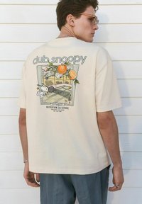 Camiseta de algodón beige con un estampado gráfico en la parte posterior, que representa una escena de tenis con Snoopy, naranjas y texto. Cuello redondo, corte relajado.