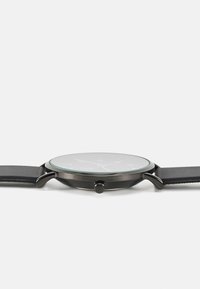 Montre avec un bracelet en cuir noir, boîtier rond en métal argenté, verre dépoli et index d'heure blancs minimalistes. Dispose d'une couronne latérale pour les réglages.