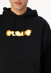 Svart hettegenser med teksturert trykk som har en oransje og hvit logo. Snorer ved halsen og en frontlomme. Mykt stoff.
