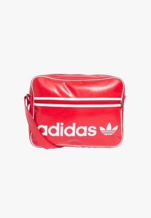 Bolsa transversal sintética vermelha com riscas diagonais brancas. Grande logótipo branco da "adidas" e gráfico do trevo na parte da frente. Compartmento principal com fecho éclair.