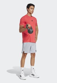 Camiseta deportiva roja con logo negro, pantalones cortos grises, kettlebell negro, calcetines blancos y zapatillas blancas con franjas negras, en pose de pie.
