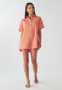 Kortärmad sparkdräkt i persikofärgat tyg, med krage, knäppning framtill och en bröstficka. Mjuk textur med avslappnad passform.