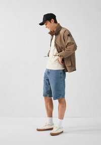 Lichtbruine zip-up jas, witte T-shirt, blauwe denim shorts, witte en tan sneakers en een zwarte pet gedragen door een persoon die zijwaarts staat.