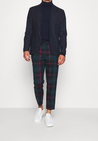 Granatowa marynarka, czarny golf, zielone spodnie w kratkę z czerwonymi i niebieskimi akcentami oraz białe sneakersy. Dopasowany krój z otwartymi kieszeniami.