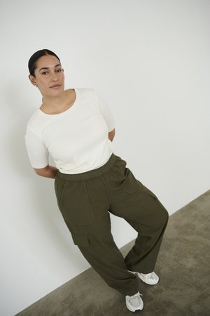 Femme aux cheveux foncés attachés, portant une chemise blanche, un pantalon cargo olive et des baskets blanches, debout devant un mur blanc uni.