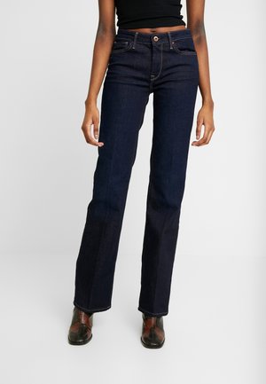 Kvinde iført mørkeblå jeans med lige ben, sort cropped top og brune slangeskindsstøvler, stående mod en ensfarvet lys baggrund.