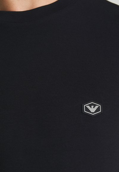 T-shirt in cotone nero con colletto girocollo e un patch logo esagonale bianco con design lineare sul lato sinistro del petto. Texture liscia.