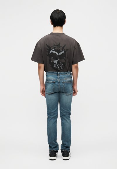 Γκρι μπλουζάκι με μαύρο γραφικό σχέδιο κρανίου, σε συνδυασμό με μπλε τζιν παντελόνι με εφαρμοστή κοπή και πίσω τσέπες. Μαύρα αθλητικά παπούτσια.