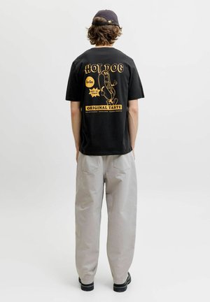 Ung person iført en sort kasket, sort t-shirt med gul hotdog-grafik og tekst, lysegrå bukser og sorte sko, stående med ryggen til.