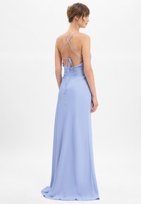 Femme portant une robe longue de couleur bleu clair avec de fines bretelles croisées et un dos nu noué en nœud, se tenant devant un fond blanc.