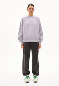 Vrouw die staat in een lichtpaarse sweatshirt met de tekst "ARMEDANGELS", donkere losse broek en witte sneakers met groene veters, voor een witte achtergrond.