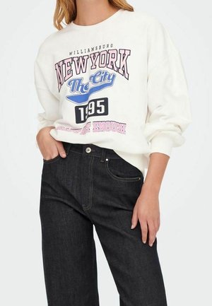 Person iført hvid sweatshirt med teksten "New York The City 1995" og sorte højtrissede jeans, hånd i lommen, mod ensfarvet baggrund.