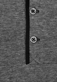 Šedé polo tričko s texturovanou tkaninou, černými akcenty a dvěma kovovými knoflíky u límce; minimalistický design s subtilním detailem loga.