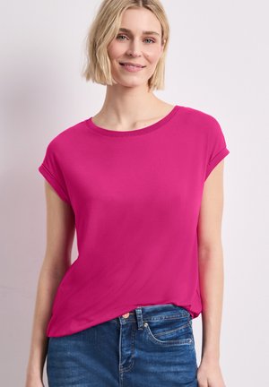 Blonde Frau mit kurzen Haaren, die ein knallpinkes kurzärmeliges Oberteil und blaue Jeans trägt, lächelnd vor einem schlichten hellen Hintergrund.