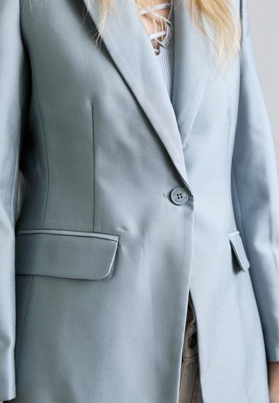 Kobieta w jasnoszarym żakiecie zapinanym na pojedynczy guzik, nosząca białą bluzkę na sznurki i beżowe spodnie, ukazana od ramion do bioder.