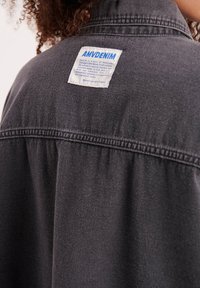 Oseba s kodrastimi lasmi, oblečena v temnosiv denim jopič z belo in modro nalepko AMVDENIM na zgornjem delu hrbta.