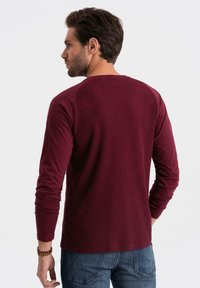 Chemise à manches longues bordeaux avec texture côtelée et manches raglan. Conçue avec une coupe confortable et des poignets fins, associée à un jean bleu.