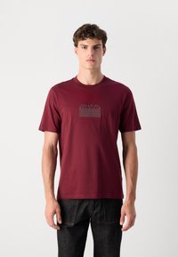 Burgundi spalvos medvilnės T-marškiniai su trumpomis rankovėmis, apvalia iškirpte ir tekstūruotu logotipu, pavaizduotu stačiakampio forma priekyje.