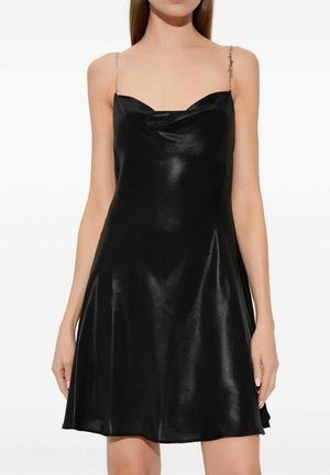 Femme portant une robe nuisette en satin noir sans manches avec de fines bretelles en chaîne et une jupe légèrement évasée, debout devant un fond blanc.