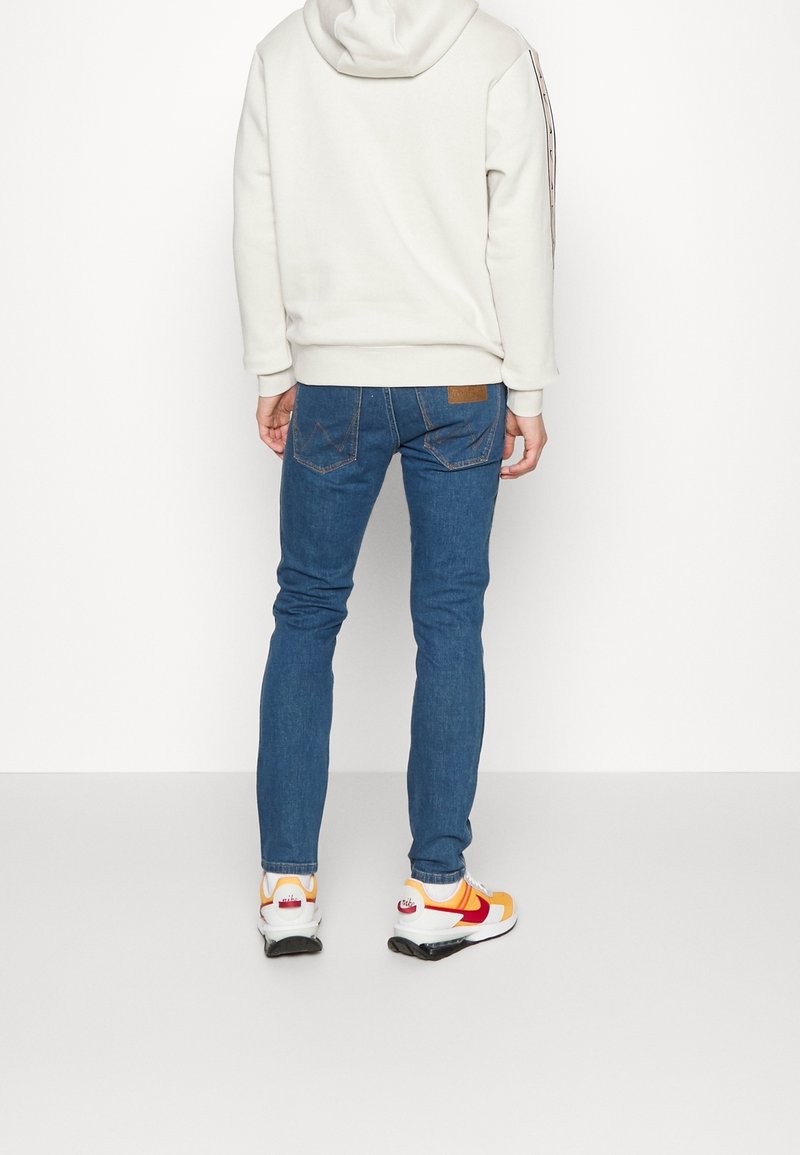 Harmaa huppari, jossa on huppu, yhdistettynä sinisiin slim-fit farkkuihin. Tennarit ovat keltainen, punainen ja valkoinen värimaailma kontrastiväreillä.