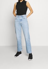 Kvinna klädd i en ärmlös svart polo-topp, ljusblå jeans med hög midja och rak ben, samt svarta sneakers, står mot en enfärgad bakgrund.