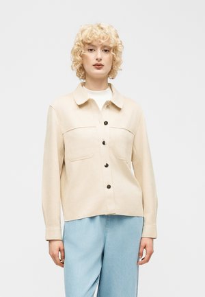 Γυναίκα με σγουρά ξανθά μαλλιά που φοράει μπεζ σακάκι με κουμπιά και τσέπες στο στήθος και ανοιχτό μπλε τζιν παντελόνι, στέκεται μπροστά σε απλό φόντο.