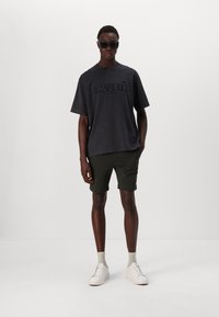 Čierne bavlnené tričko s embosovaným logom "LACOSTE", skombinované so zelenými šortkami. Model nosí slnečné okuliare a biele tenisky s béžovými ponožkami.