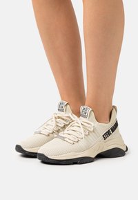 Baskets beiges en mesh avec une tige texturée, des lacets plats et un logo accent noir sur le côté. Elles disposent d'une semelle rembourrée pour le confort.