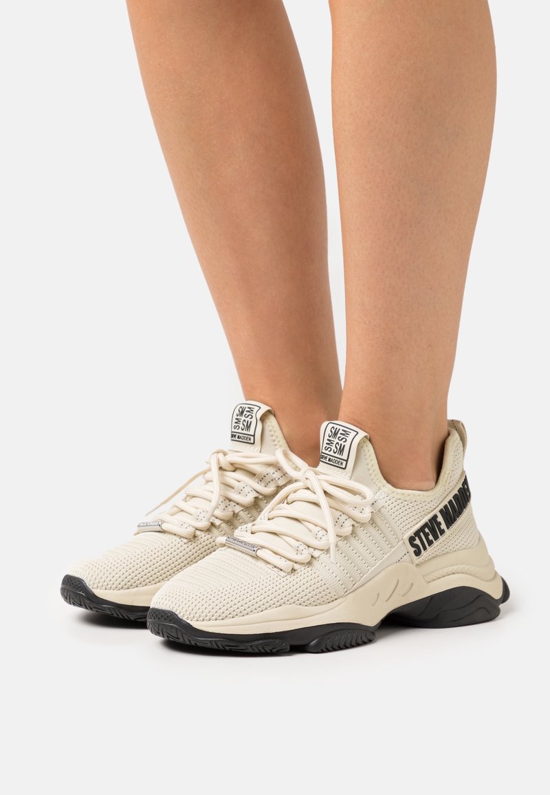 Baskets beiges en mesh avec une tige texturée, des lacets plats et un logo accent noir sur le côté. Elles disposent d'une semelle rembourrée pour le confort.