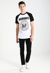 Camiseta raglán blanca y negra con el texto "Ramones" y un gráfico de un águila, combinada con pantalones negros ajustados y zapatillas blancas.