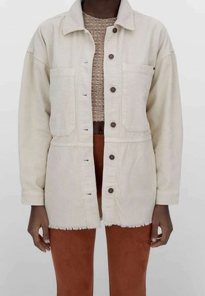 Person wearing an off-white corduroy button-up jacket over a beige textured top and rust-colored pants, standing against a plain light background.