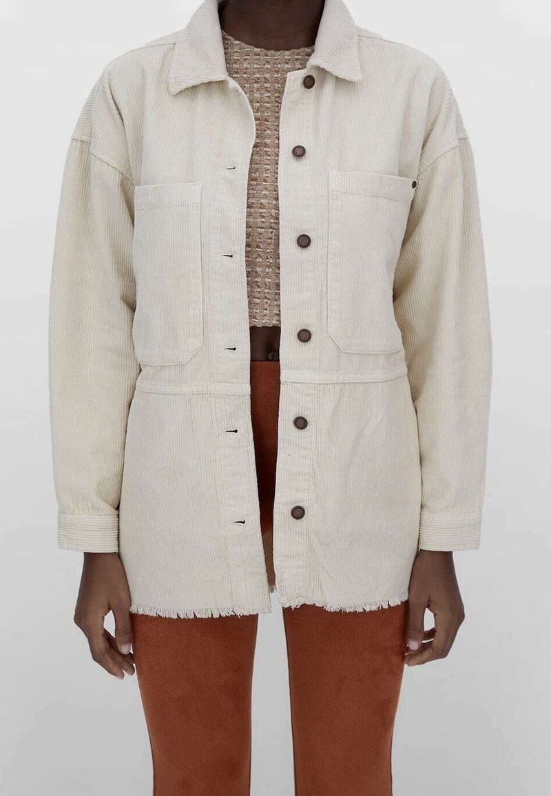 Personne portant une veste en velours côtelé écru à boutons, sur un haut texturé beige et un pantalon couleur rouille, debout devant un fond clair uni.