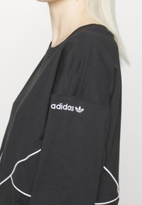 Person wearing a black Adidas sweatshirt with white piping, showing the Adidas logo on the upper sleeve against a plain background.