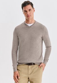 Beżowy sweter w serek wykonany z miękkiego, teksturowanego materiału z długimi rękawami. W połączeniu z jasnymi spodniami i brązowym paskiem.