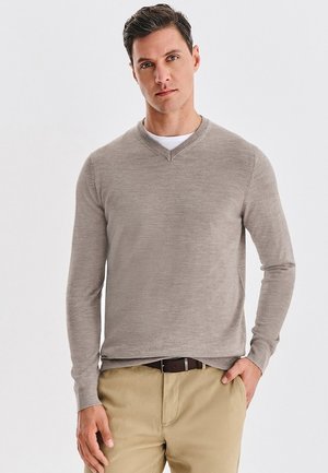 Beżowy sweter w serek wykonany z miękkiego, teksturowanego materiału z długimi rękawami. W połączeniu z jasnymi spodniami i brązowym paskiem.