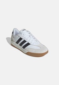 Zapatilla blanca de Adidas con rayas negras, puntera de ante beige, suela de goma y cordones blancos, presentada en ángulo para mostrar el lado exterior y la vista superior.