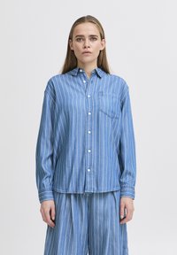 Camicia azzurra chiara, con bottoni, a righe verticali bianche, maniche lunghe e taschino sul petto. Realizzata in denim con una vestibilità rilassata.