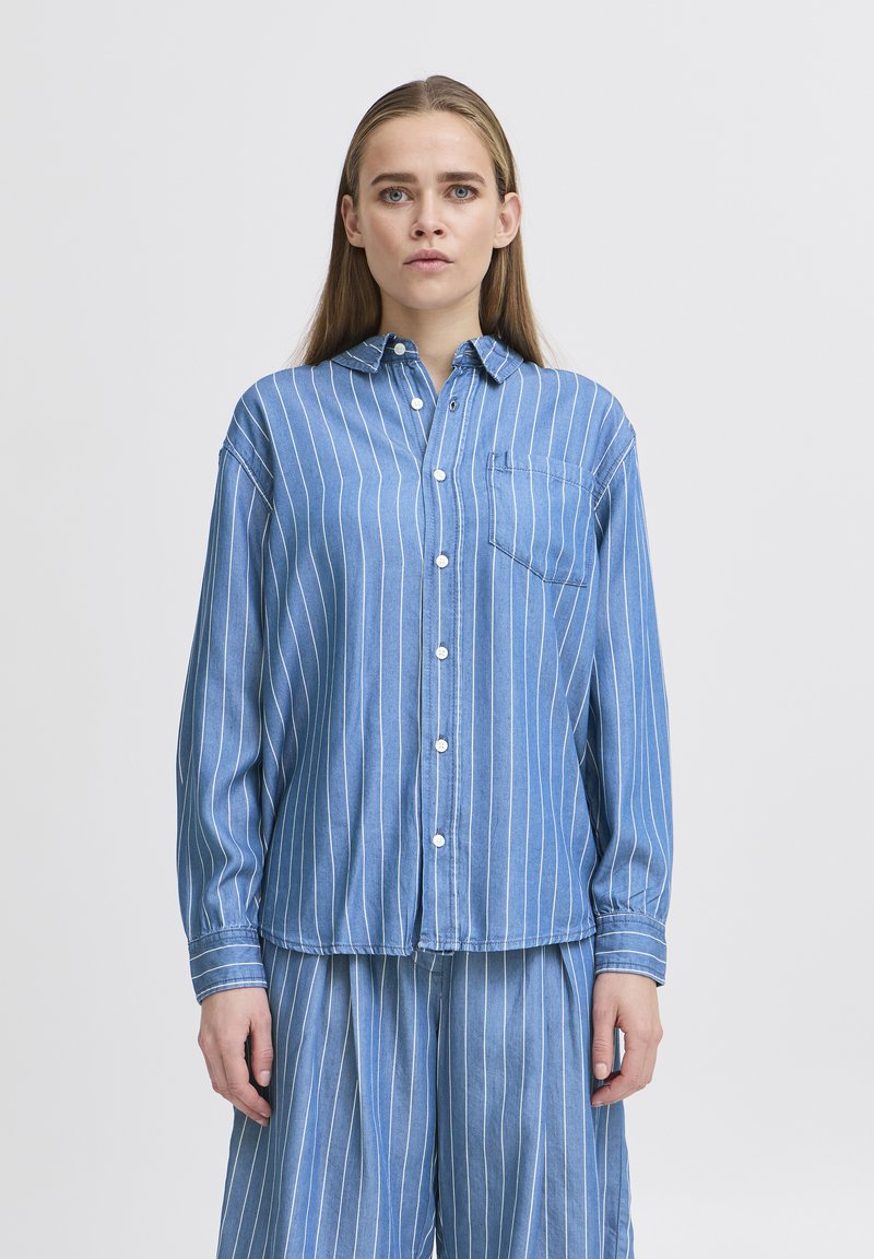Camicia azzurra chiara, con bottoni, a righe verticali bianche, maniche lunghe e taschino sul petto. Realizzata in denim con una vestibilità rilassata.
