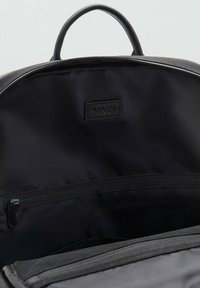 Interior de mochila negra con un forro de tela suave, que cuenta con un bolsillo con cremallera y una etiqueta grabada con "MNG". Diseño minimalista.