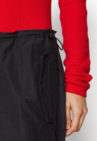 Pantalon de sport noir avec taille élastique et cordon de serrage. Comprend une poche latérale et une texture lisse, associé à un haut à manches longues rouge côtelé.