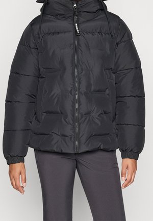 Personne portant une doudoune noire avec une fermeture éclair frontale et des poignets élastiques, portée avec un pantalon gris foncé sur un fond uni.