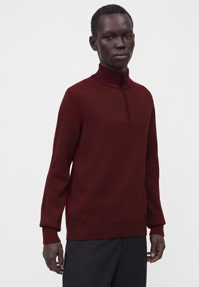 Homme portant un pull en maille bordeaux avec un col à demi-fermeture éclair et un pantalon noir, debout devant un fond clair uni.