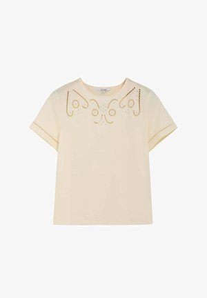 Haut crème à manches courtes avec encolure ronde, orné d'embellissements à clous dorés et argentés en motifs tourbillonnants symétriques sur la poitrine et les bords des manches.