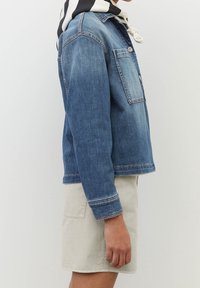 Giacca di jeans in blu chiaro con chiusura a bottoni, tasca sul petto e cuciture in contrasto; abbinata a un completo di pantaloni corti di colore chiaro.