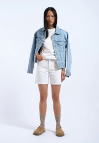 Chaqueta de mezclilla azul claro sobre una camiseta blanca, combinada con pantalones cortos blancos. Zuecos de ante beige usados con calcetines marrones de diseño. Diseño sencillo.