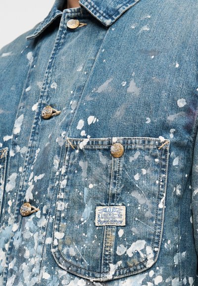 Τζιν μπουφάν σε ανοιχτό μπλε χρώμα με πιτσιλιές λευκής βαφής, με χρυσά κουμπιά, δύο εμπρόσθιες τσέπες και υφασμένη ετικέτα στην δεξιά τσέπη.