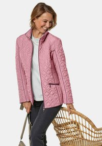 Gequilted roze jack met een hoge kraag, ritszakken en contrasterende bies. Draagt het over een wit topje, gecombineerd met donkere broeken, en houdt een geweven mand vast.